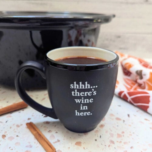 A mug filled with Slow Cooker Warm Apple Cinnamon Spiced Wine with a slow cooker behind it.