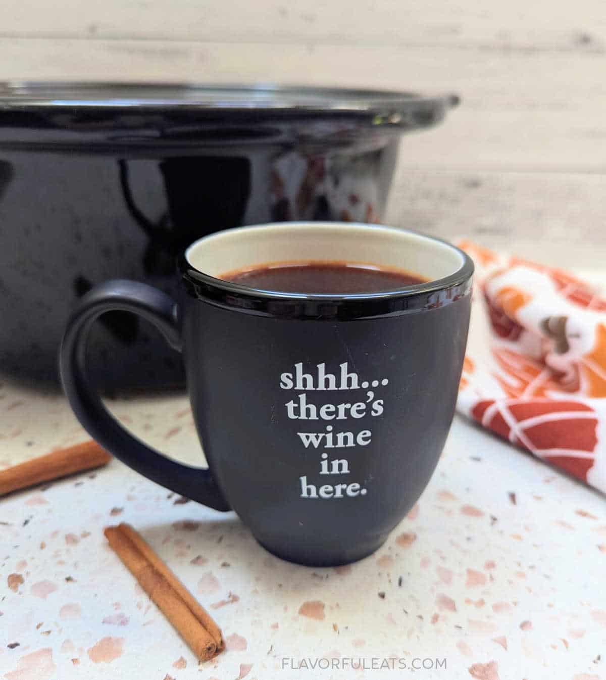 A mug filled with Slow Cooker Warm Apple Cinnamon Spiced Wine with a slow cooker behind it.