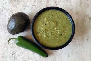 Roasted Tomatillo Avocado Salsa in a bowl with a jalapeno and avocado beside it.