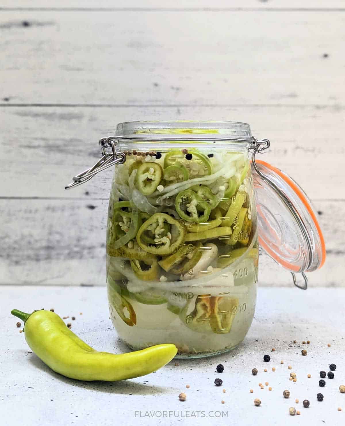 A photo of Hot Banana Peppers & Onions in a jar with a fresh hot banana pepper sitting beside it.