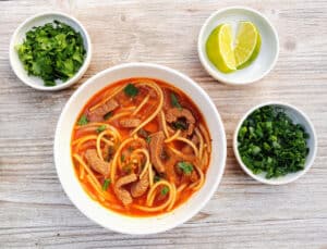 Spicy Thai Beef Noodle Soup in a bowl with small bowls of limes, cilantro, and scallions beside it.