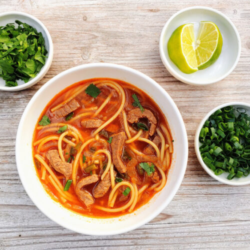 Spicy Thai Beef Noodle Soup in a bowl with small bowls of limes, cilantro, and scallions beside it.
