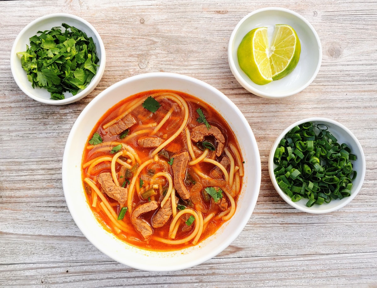 Spicy Thai Beef Noodle Soup in a bowl with small bowls of limes, cilantro, and scallions beside it.