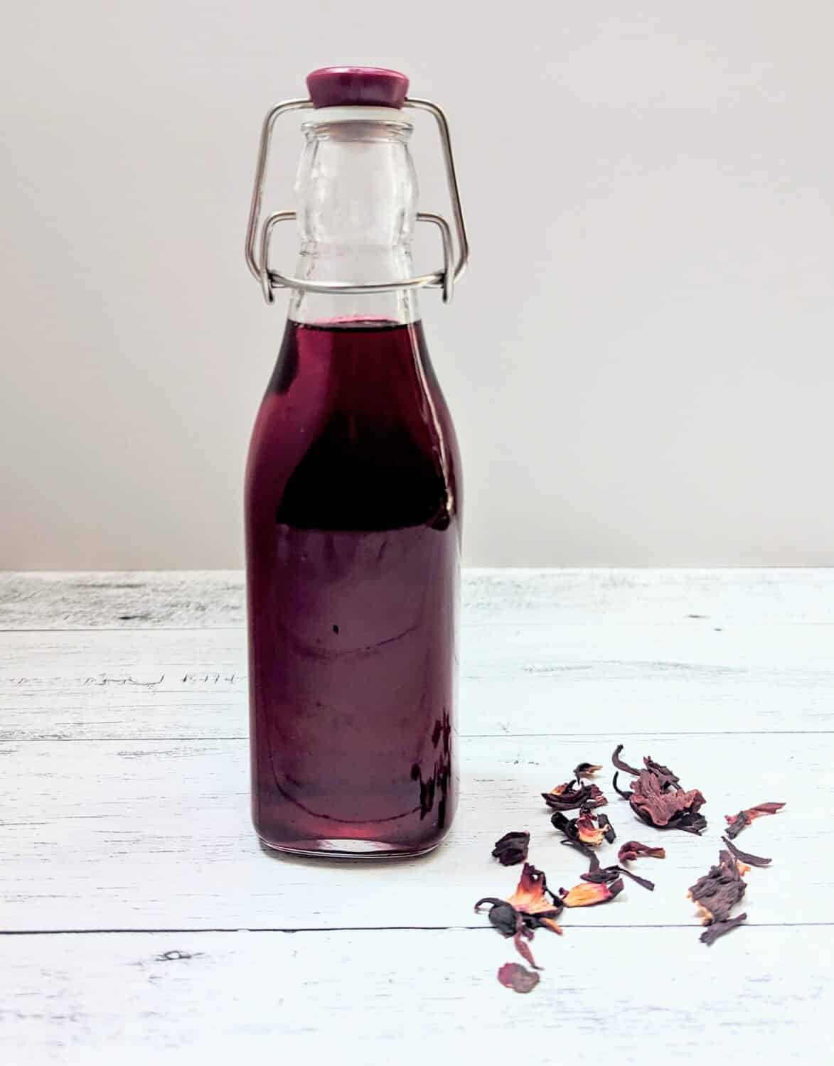 A swing-top bottle filled with Hibiscus Simple Syrup with dried hibiscus flowers beside it.