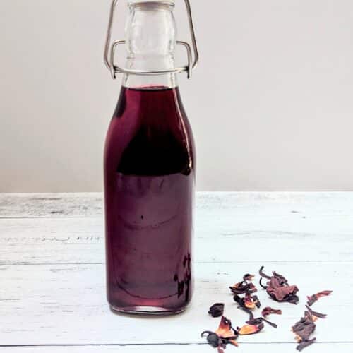 A swing-top bottle filled with Hibiscus Simple Syrup with dried hibiscus flowers beside it.