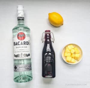 Ingredients needed to make a Hawaiian Smash Martini laid out - white rum, hibiscus simple syrup, lemon, and pineapple.