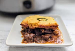 A serving of Slow Cooker BBQ Beef Cornbread Casserole on a white plate with a slow cooker in the background.