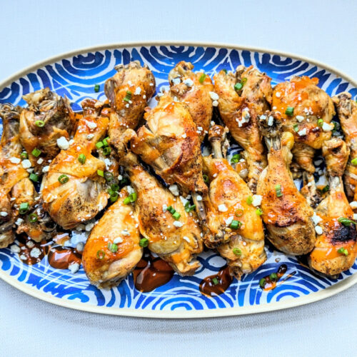 A blue platter with Slow Cooker Buffalo Chicken Drumsticks stacked on it with sliced scallions and blue cheese crumbles garnished on top