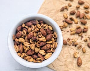 A small white bowl full of Spicy Curry Roasted Nuts with some nuts on a brown piece of parchment paper.