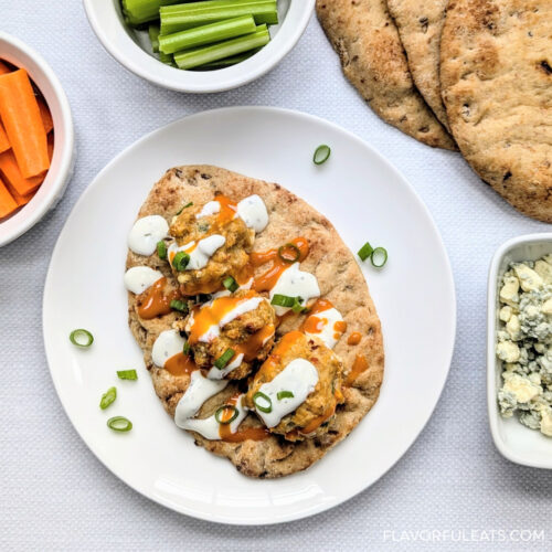 Baked Buffalo Chicken Meatballs on a naan with condiments on top beside a stack of naan and bowls of carrots and celery.