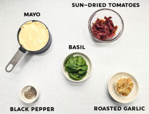 A photo of ingredients needed to make Roasted Garlic Sun-Dried Tomato Aioli, which includes mayo, sun-dried tomatoes, basil, pepper, and roasted garlic.
