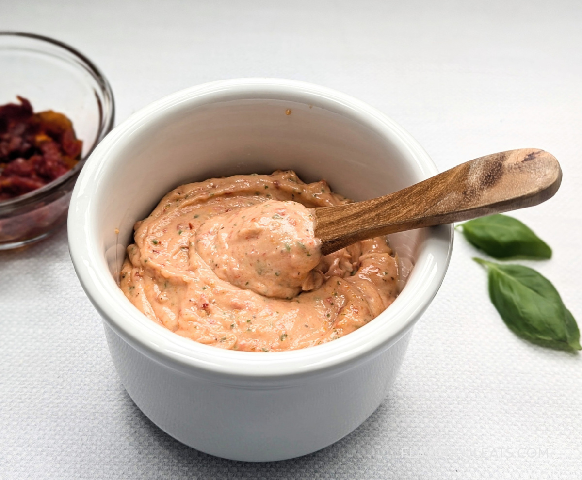 A white bowl with Roasted Garlic Sun-Dried Tomato Aioli with a spoon dipping in it.