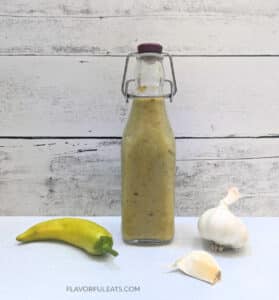 A glass bottle with Hot Banana Pepper Garlic Hot Sauce with garlic and a banana pepper beside it.
