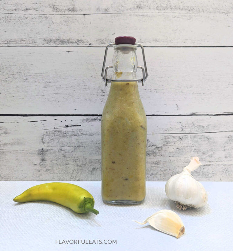 A glass bottle with Hot Banana Pepper Garlic Hot Sauce with garlic and a banana pepper beside it.