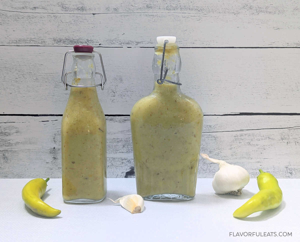 Two bottles of Hot Banana Pepper Garlic Hot Sauce with garlic and banana peppers beside them.