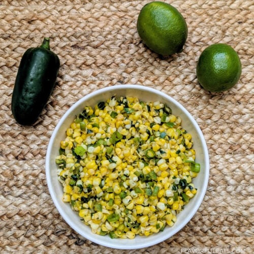 Slow Cooker Jalapeno Lime Corn in a white bowl on a placemat with limes and a jalapeno beside it.